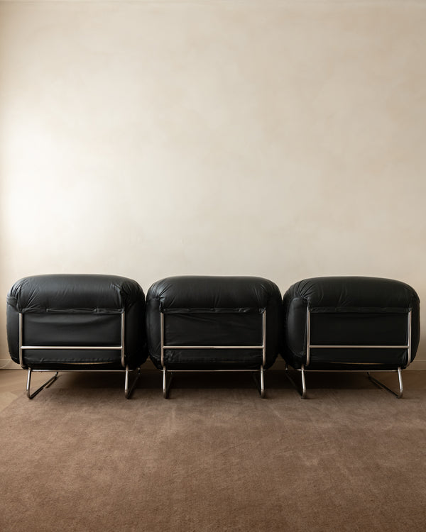 Three matching black leather lounge chairs with chrome legs arranged side-by-side against a warm beige wall and brown carpet.