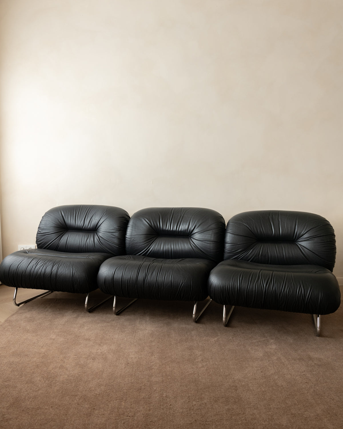 A three-seat black leather sofa with a tufted backrest and slim metal legs, positioned against a plain beige wall on a brown carpeted floor.