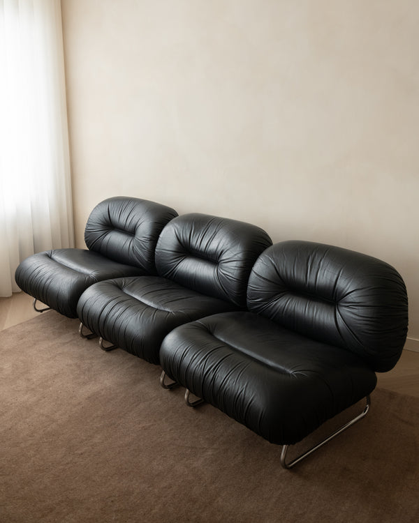 A three-seater modular sofa upholstered in black leather, featuring a low, rounded, tufted design with chrome legs, set against a neutral beige wall and carpeted floor.