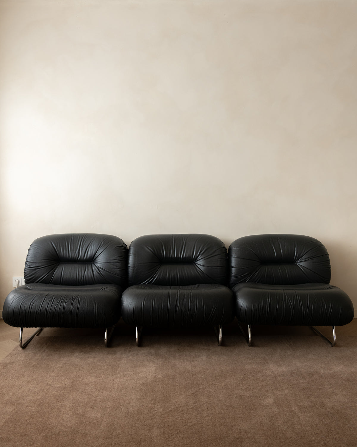 A three-seat black leather sofa with a modern curved design and slim metal legs, set against a plain beige wall and brown carpet.