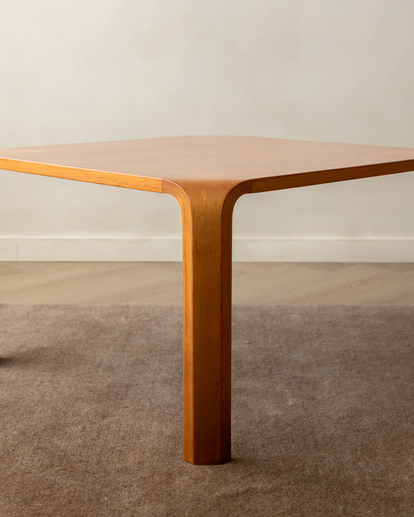 A mid-century modern wooden table with a hexagonal top and a curved, tapered pedestal leg, shown in a warm medium-brown finish against a neutral beige wall and carpet.