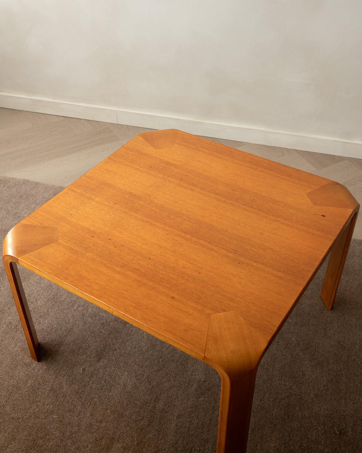A square wooden side table with rounded corners and curved legs, shown in a room setting on a gray carpet against a white wall.