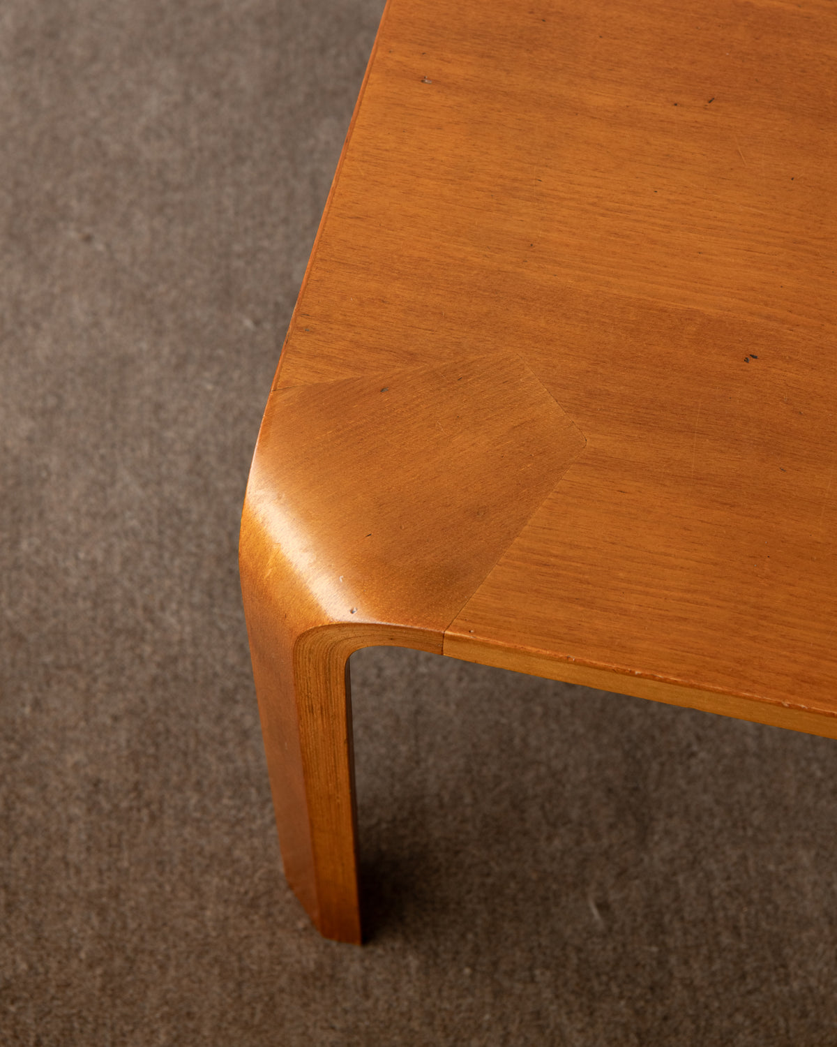 Close-up of a curved wooden table corner with a warm honey-toned wood finish, photographed on a gray carpeted floor.