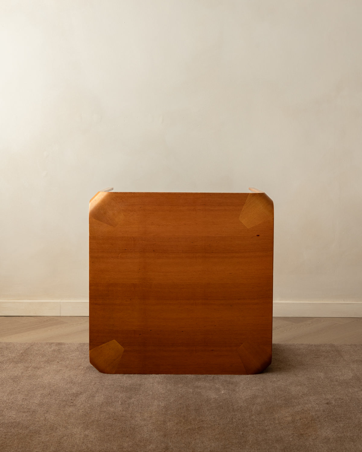 A square wooden tabletop or desk top with rounded corners and visible wood grain, photographed standing upright against a plain beige wall on a carpeted floor.