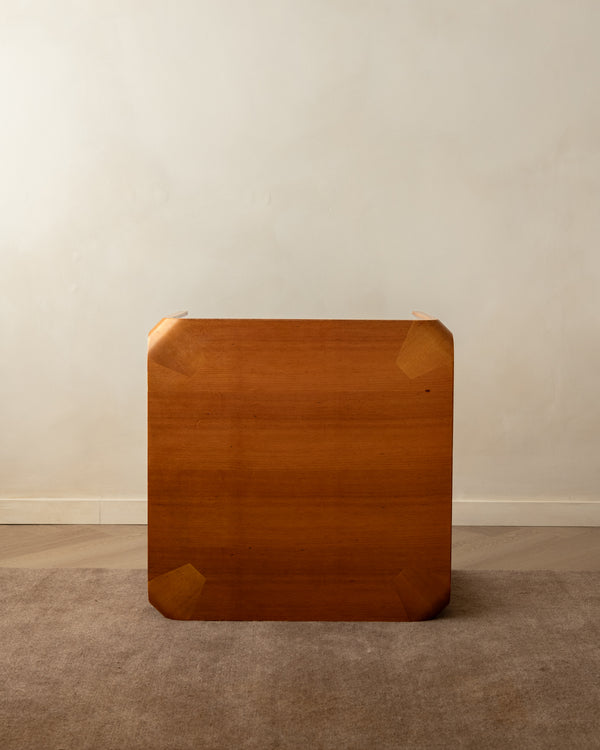A square wooden tabletop or desk top with rounded corners and visible wood grain, photographed standing upright against a plain beige wall on a carpeted floor.