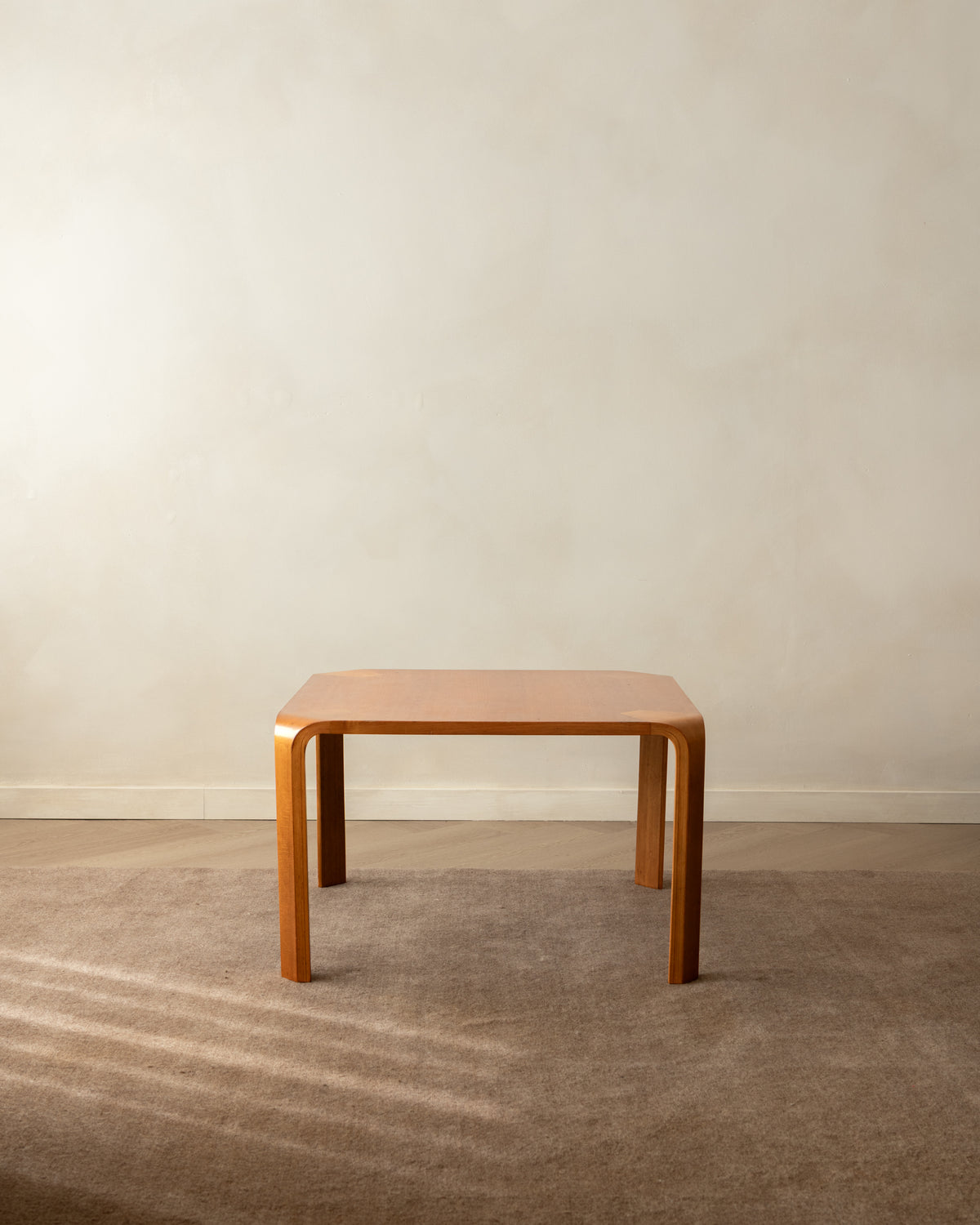 A square wooden table with a warm orange-brown finish and gently curved legs, set on a textured beige carpet against a neutral beige wall.
