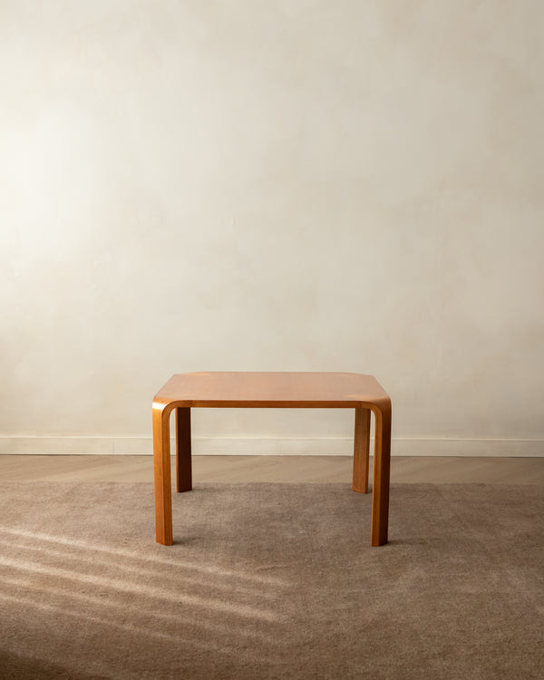 A square wooden table with a warm orange-brown finish and gently curved legs, set on a textured beige carpet against a neutral beige wall.